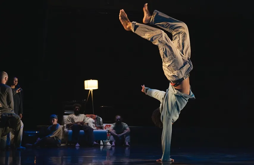 A person does a one handed handstand in the foreground while a couch full of people watch from the background.